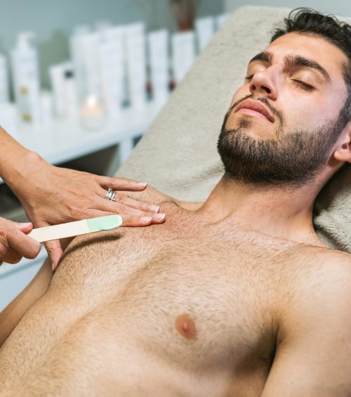 Unrecognizable cosmetologist appliyng wax hair on chest of relaxed shirtless male client lying on bed in beauty salon
